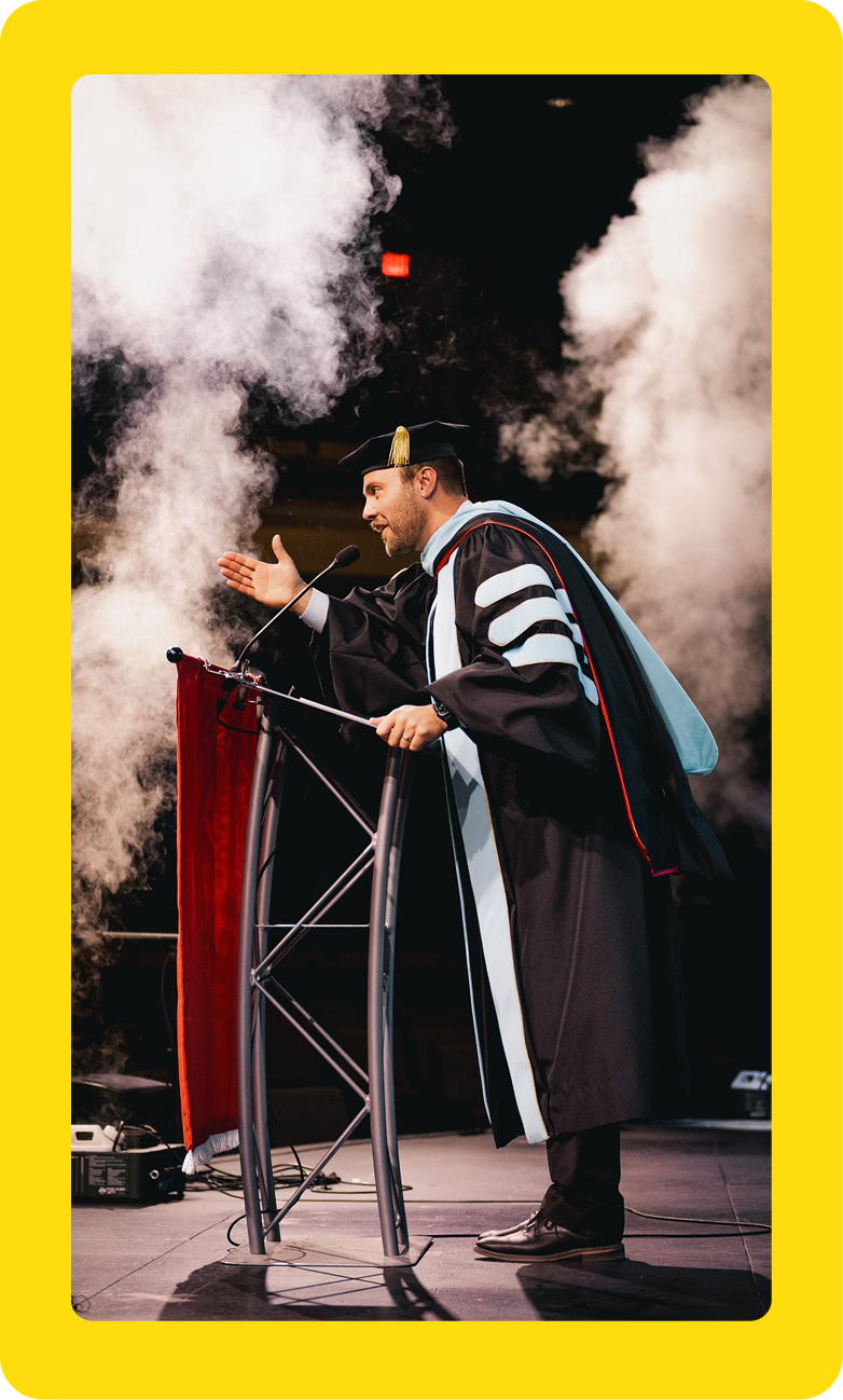Speaker at podium on stage with smoke effects, wearing a black robe with white stripes and a tall hat