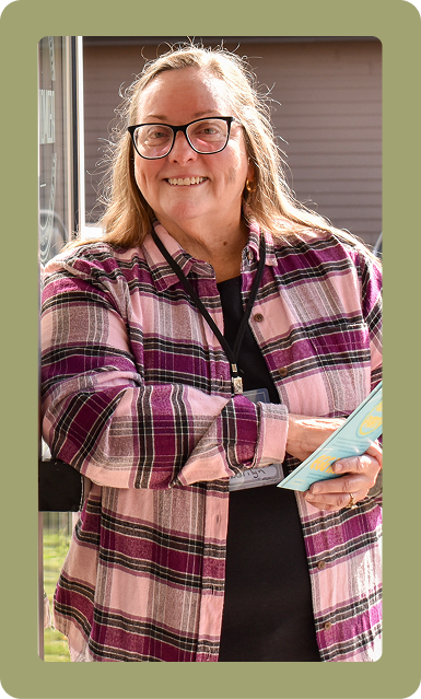 Smiling person in glasses and pink plaid shirt holding a book outdoors.