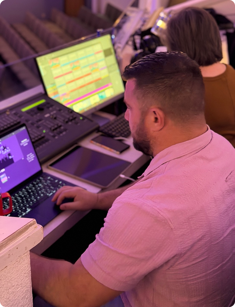 Person operating audio controls at a mixing console with lit monitors in a purple-lit studio.
