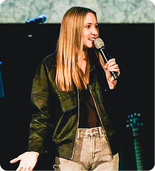 Woman speaking into a microphone on stage, wearing a dark jacket and light jeans.