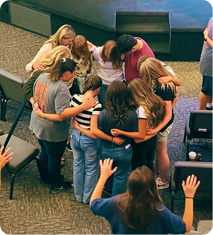 Group of people standing in a circle with arms around each other in a carpeted room