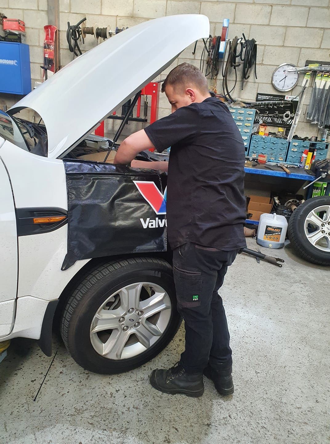 Mechanic working on a white car with the hood open in a garage, using a wrench.