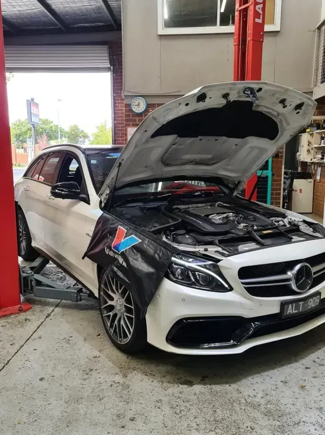 White Mercedes wagon on a lift with hood open in a garage, undergoing repair.