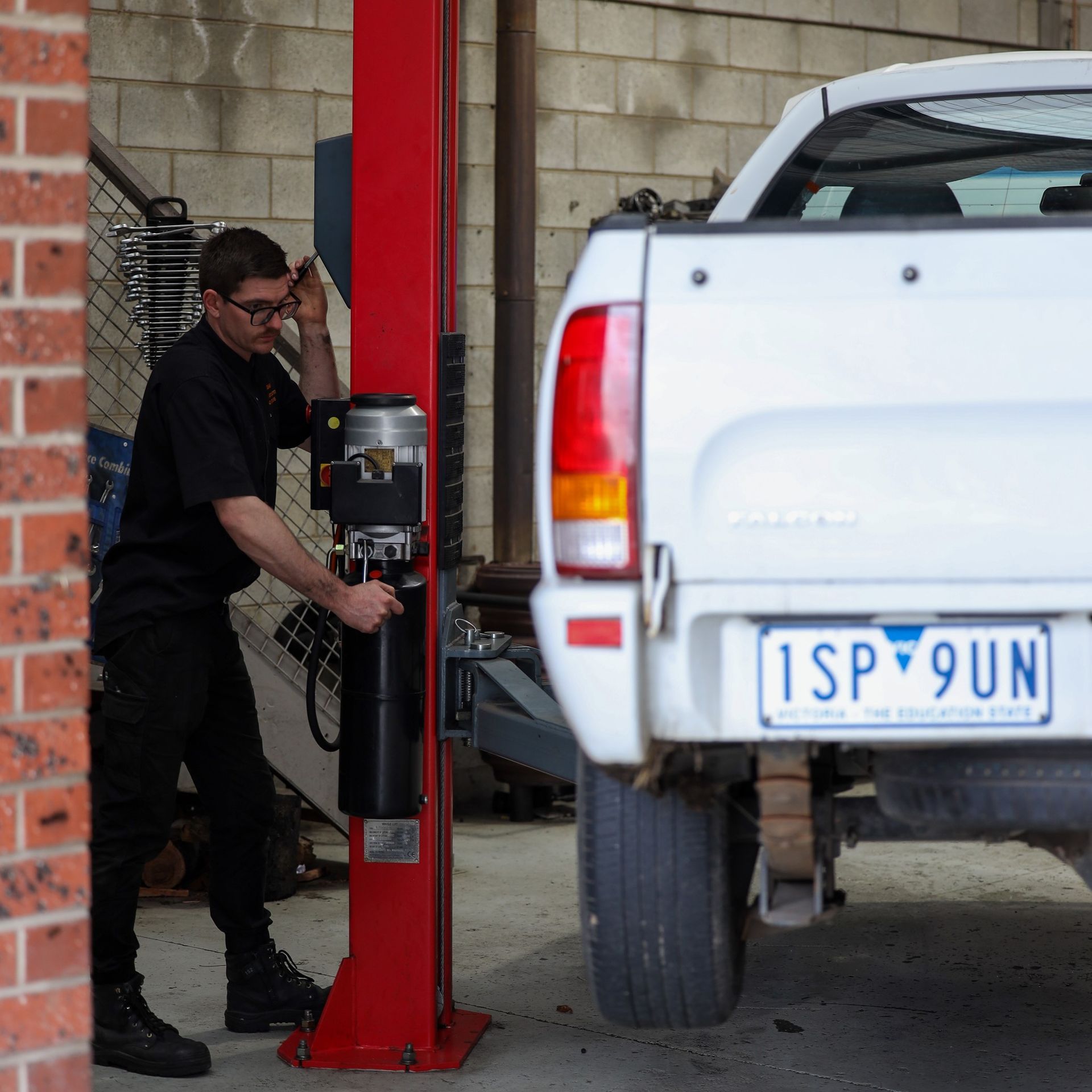 Mechanic adjusts a vehicle lift. A white pickup truck is nearby. Workshop setting, brick wall. — COOTA Automotive Services in Ballarat East, VIC