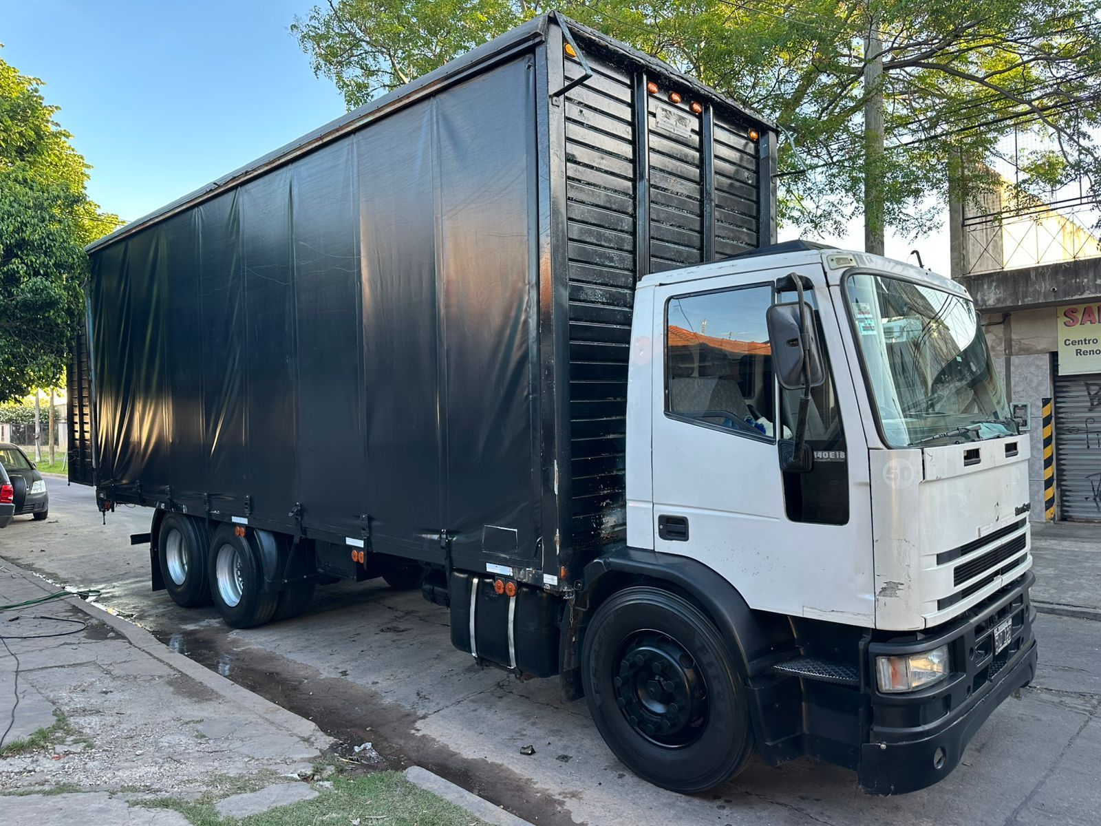 Camión de carga Ford blanco estacionado en una calle con una lona negra cubriendo el área de carga.