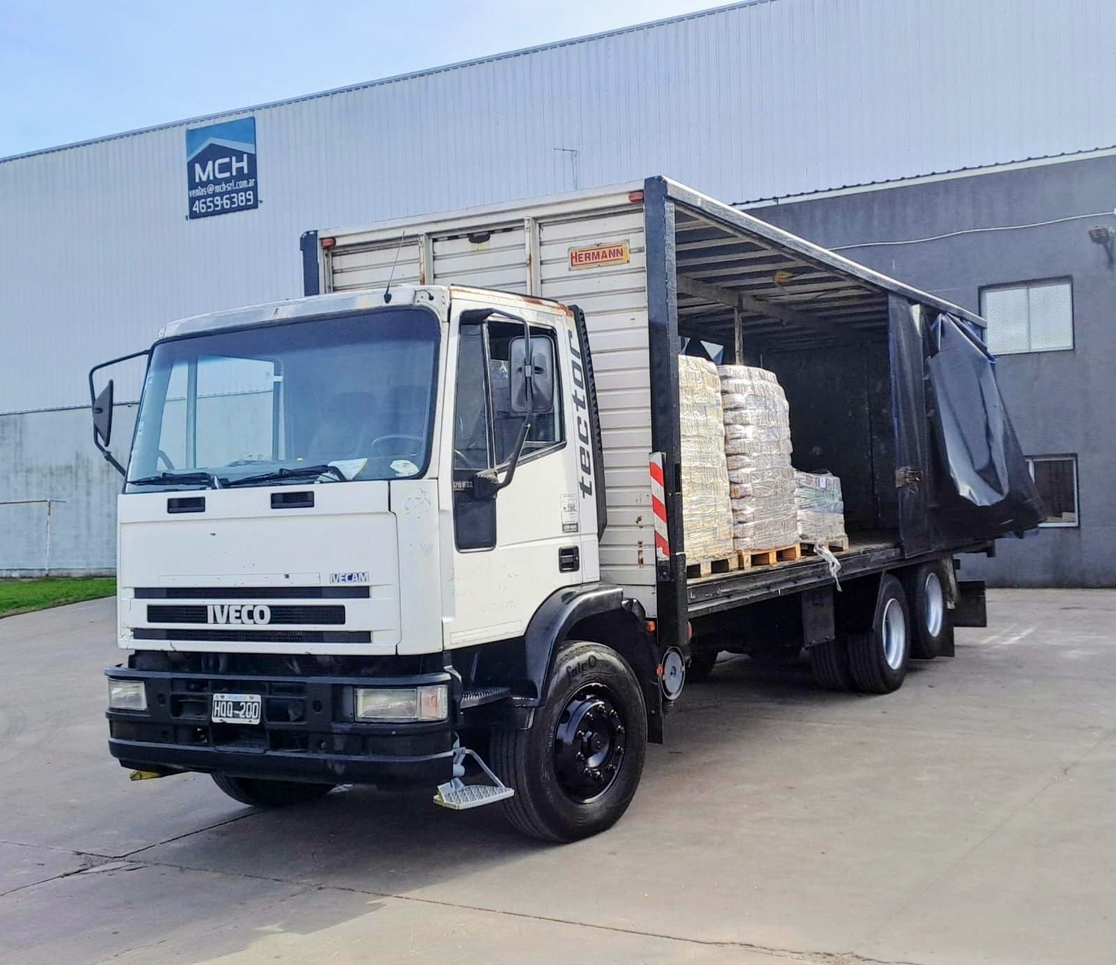 Camión Iveco blanco con zona de carga abierta cargado con pallets de ladrillos, estacionado afuera de un almacén.