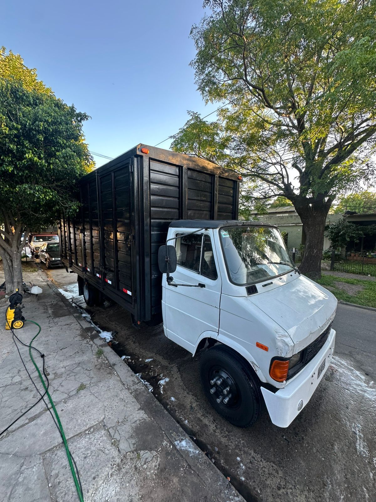 Camión de reparto blanco y negro estacionado en la acera de una calle con una plataforma de carga de madera.