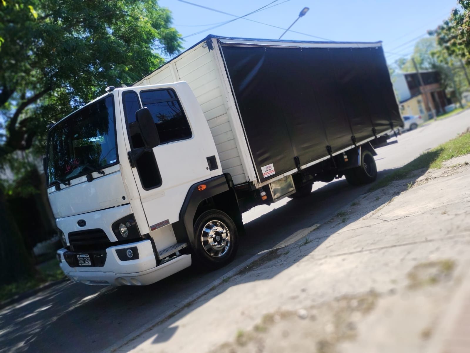 Camión de caja blanca con lona negra estacionado en una calle en pendiente, día soleado.