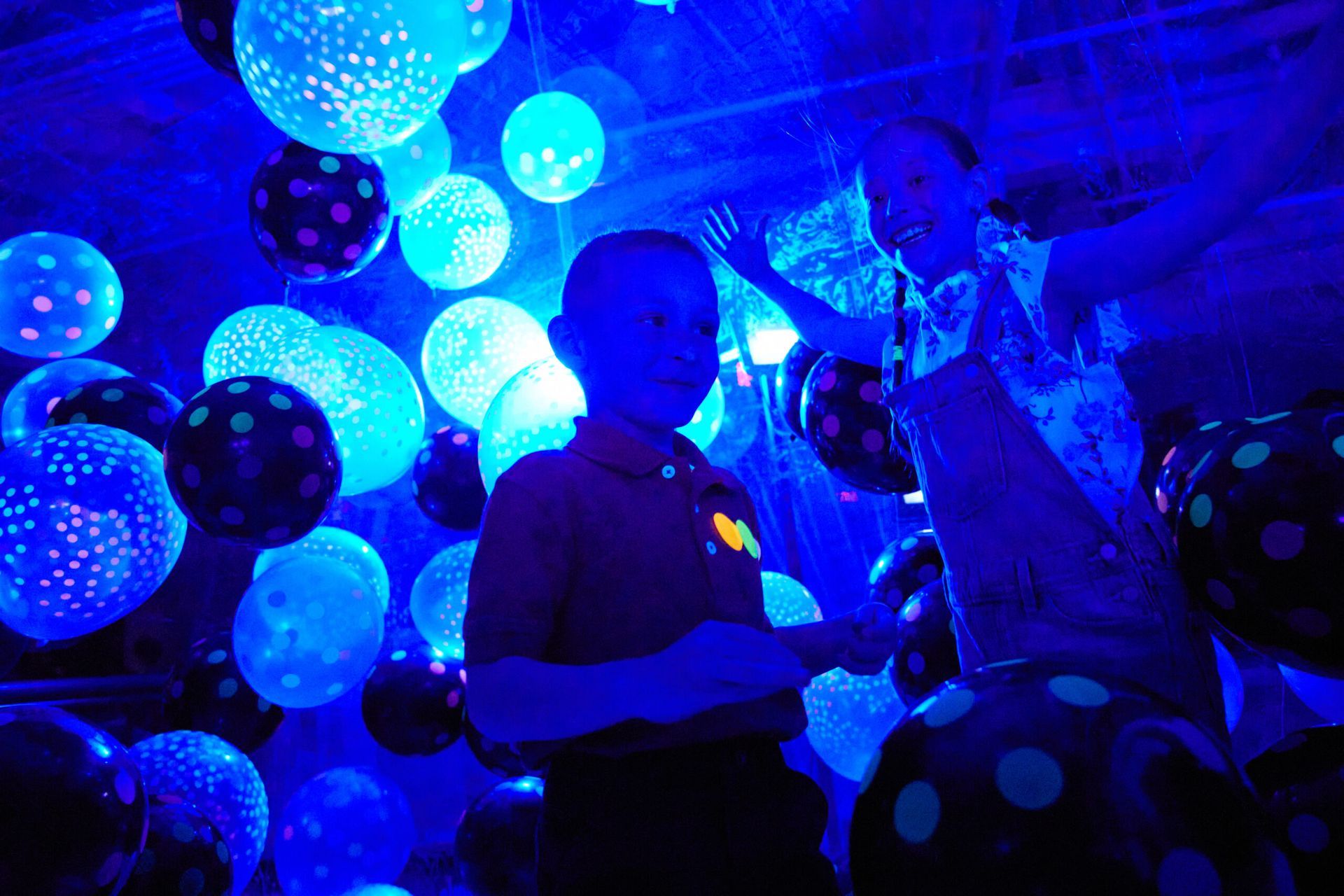Two children in a room filled with glowing blue balloons, some with polka dots.