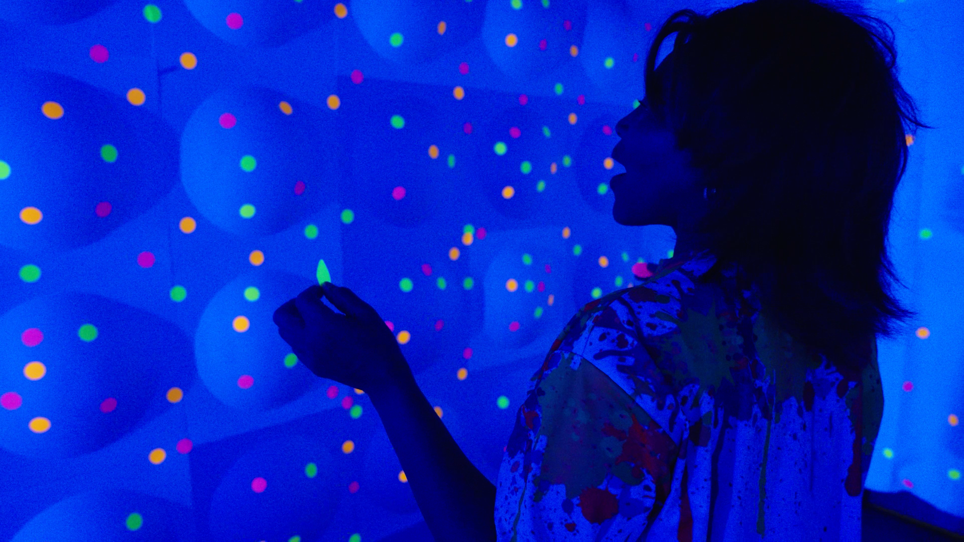 Woman in a black silhouette reaches toward glowing balloons with colorful dots in a blue-lit room.