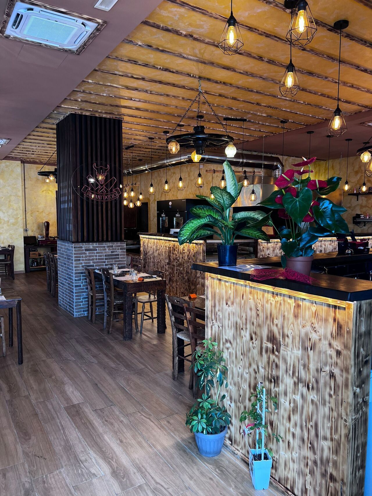 Interni del ristorante: pavimenti in legno, tavoli, bar, luci calde, piante.