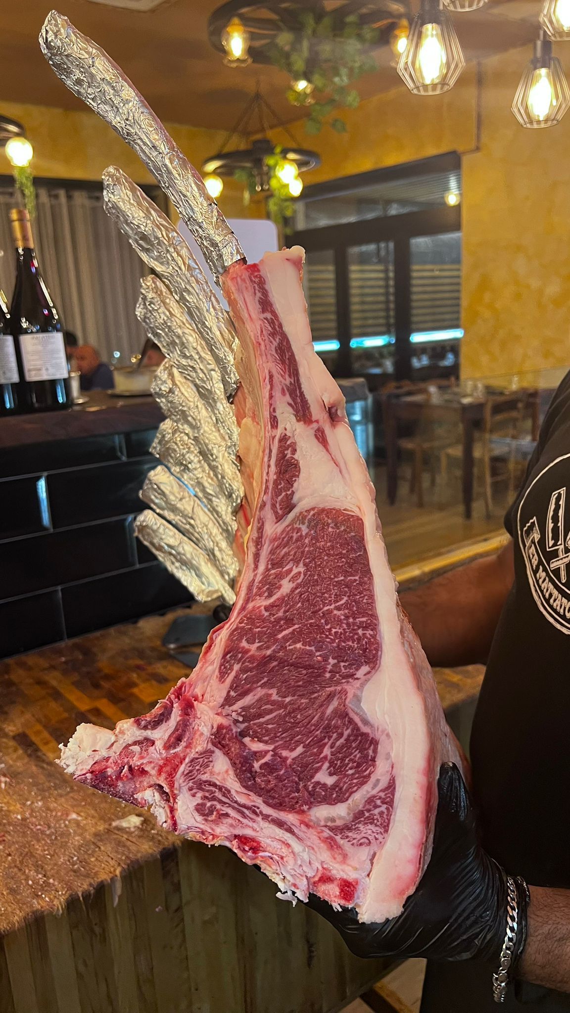 Una grande bistecca di ribeye cruda con l'osso, tenuta da una mano guantata in un ambiente da ristorante.