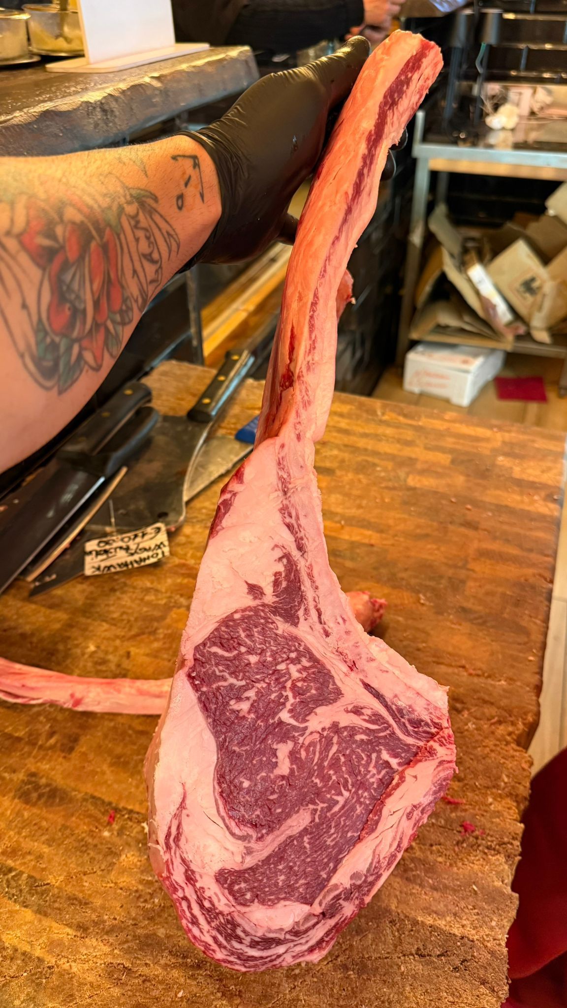 Un macellaio tiene in mano una grande bistecca di tomahawk cruda, con le venature marmoree, appoggiata su una superficie di legno.