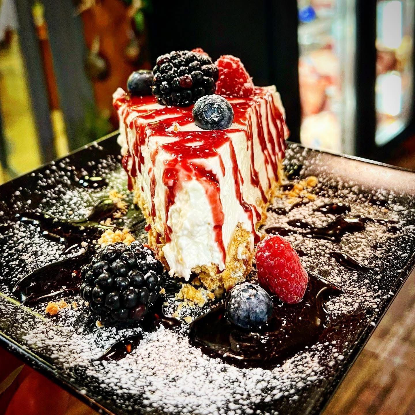 Fetta di cheesecake con guarnizione di frutti di bosco, su un piatto nero, spolverata con zucchero a velo.