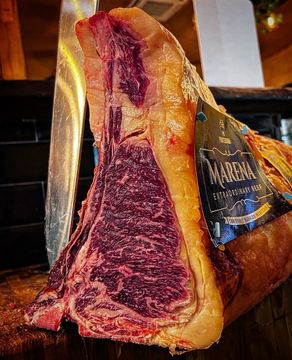 Una grande bistecca di ribeye cruda, ricca di marezzature e con osso. Accanto ad essa è posizionato un coltello.