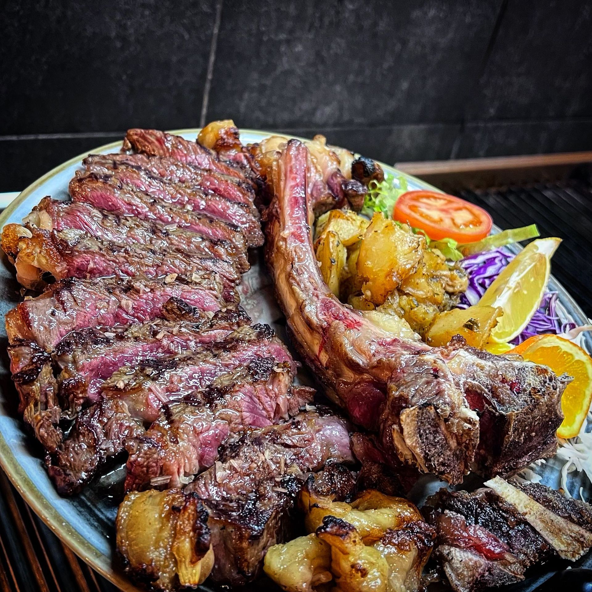 Bistecca alla griglia tagliata a fette con osso, patate, fetta d'arancia e pomodoro su un piatto.
