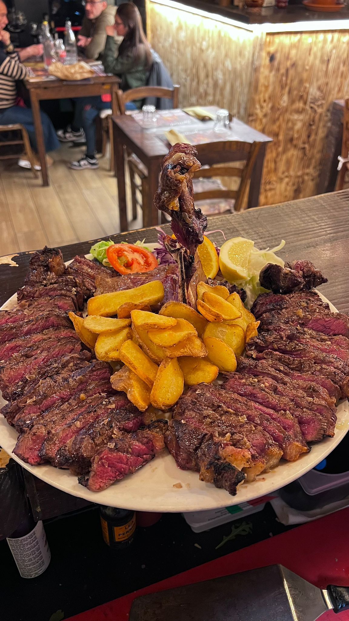 Grande piatto di bistecca affettata, patatine fritte e contorno in un ristorante. Clienti sullo sfondo.