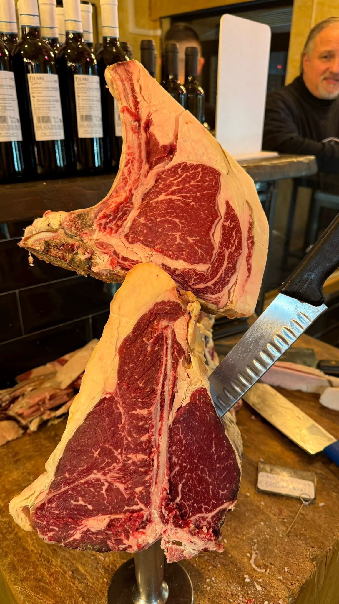 Grandi bistecche di manzo crude su un supporto con un coltello, in un ambiente da ristorante, con un uomo sorridente sullo sfondo.
