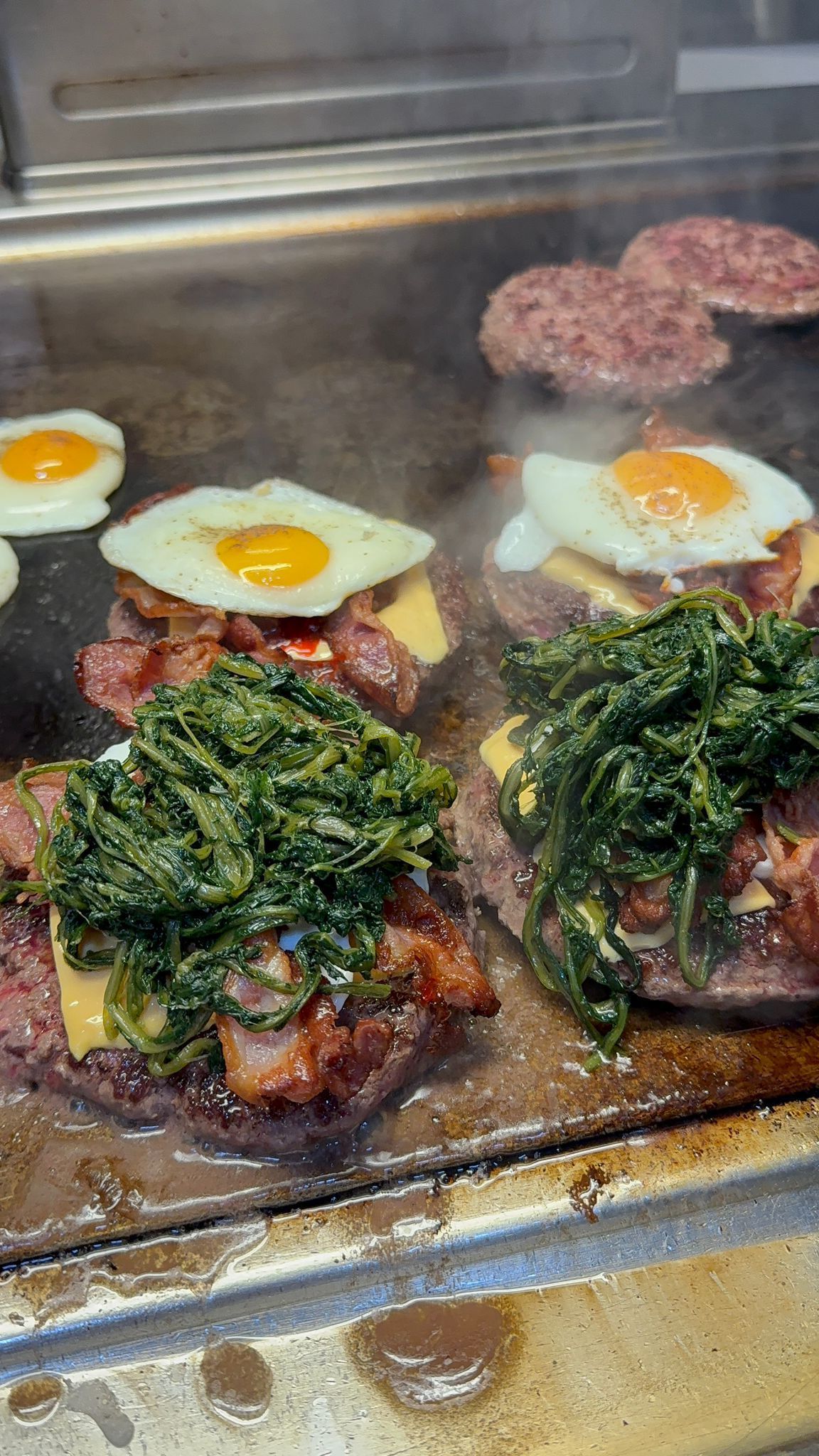 Hamburger cotti su una griglia piatta, conditi con pancetta, spinaci, formaggio e uova fritte.