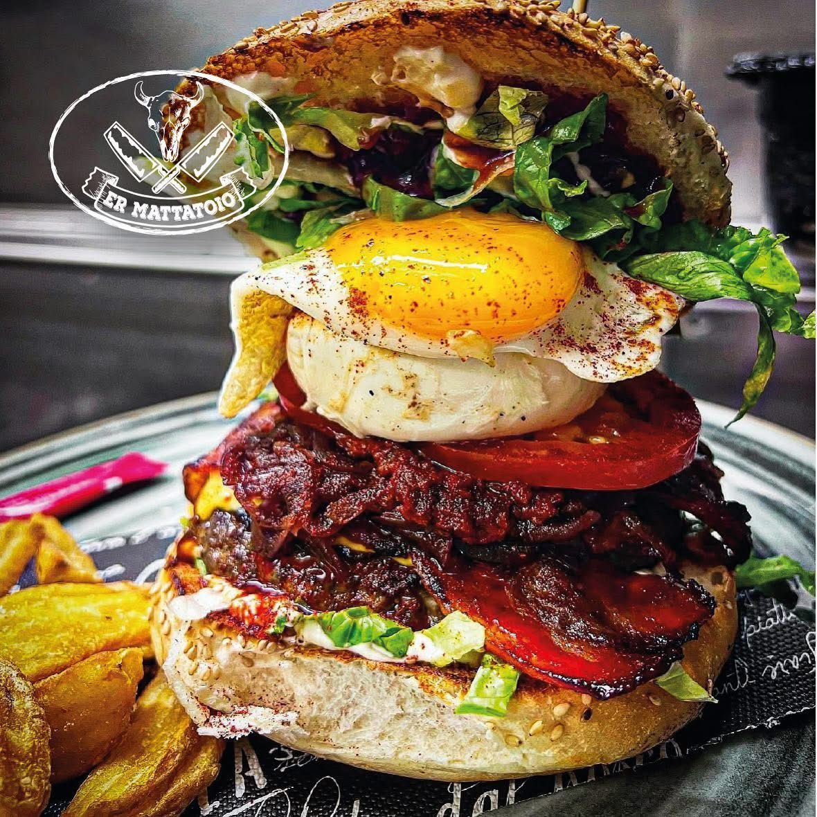 Hamburger con uovo fritto, pomodoro, pancetta e lattuga in un panino, servito con spicchi di patate.