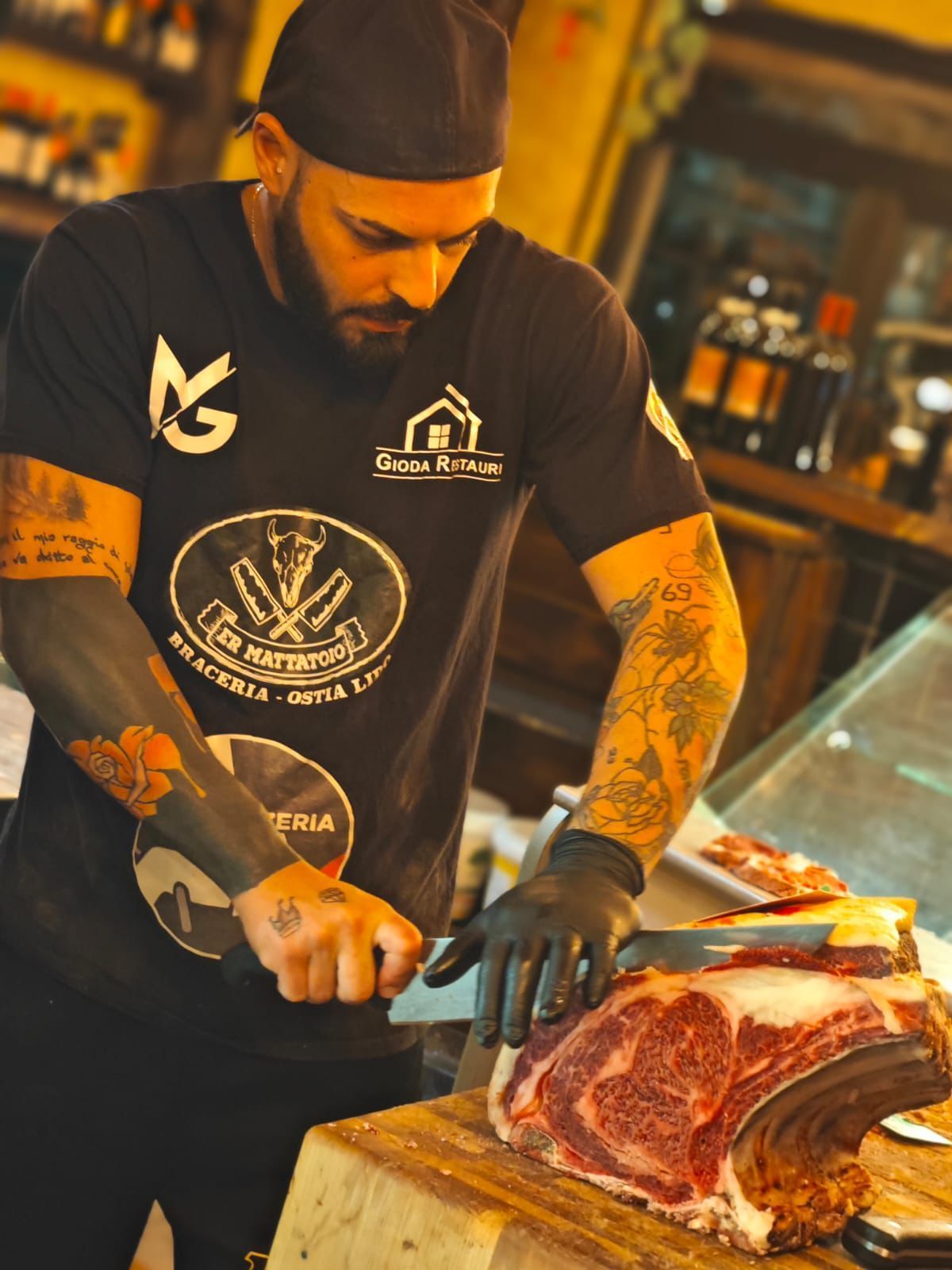 Uno chef tatuato, con una camicia nera, taglia una grande bistecca di ribeye con un coltello in un ristorante.