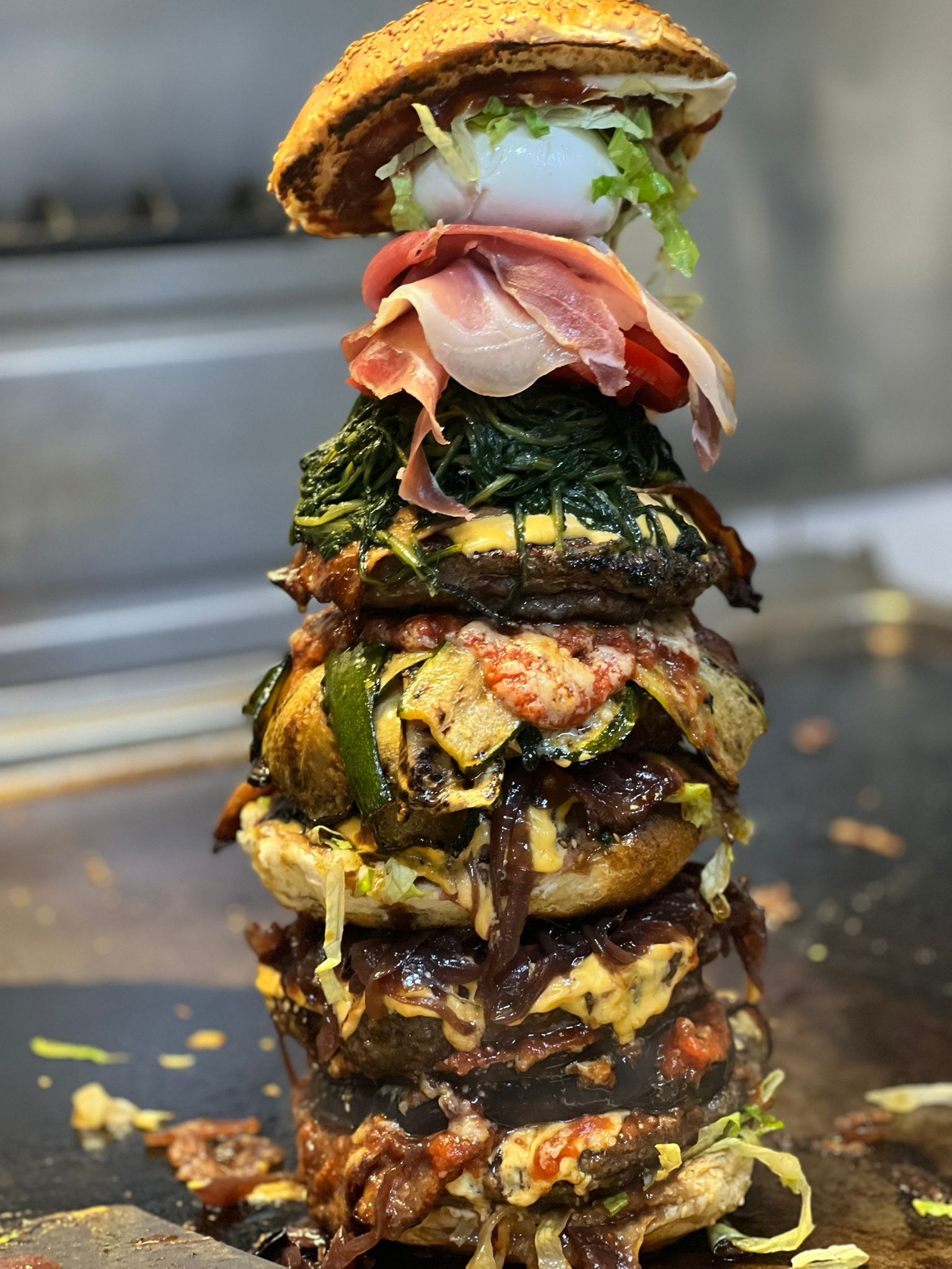Hamburger gigante con più strati, tra cui carne, verdure e condimenti, cotto sulla griglia.