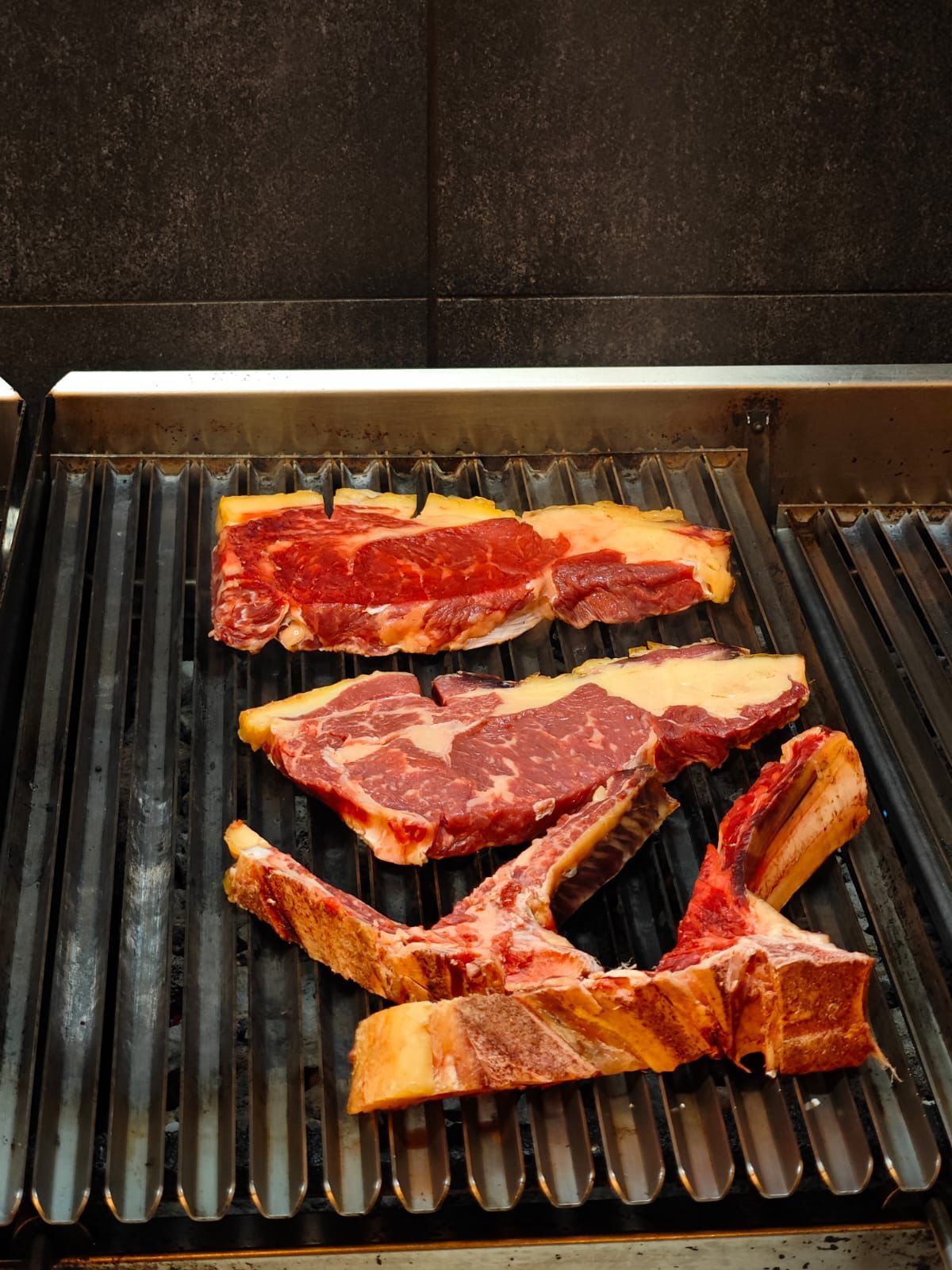 Tre bistecche tagliate spesse su una griglia calda, con segni di grigliatura visibili e sfondo scuro.