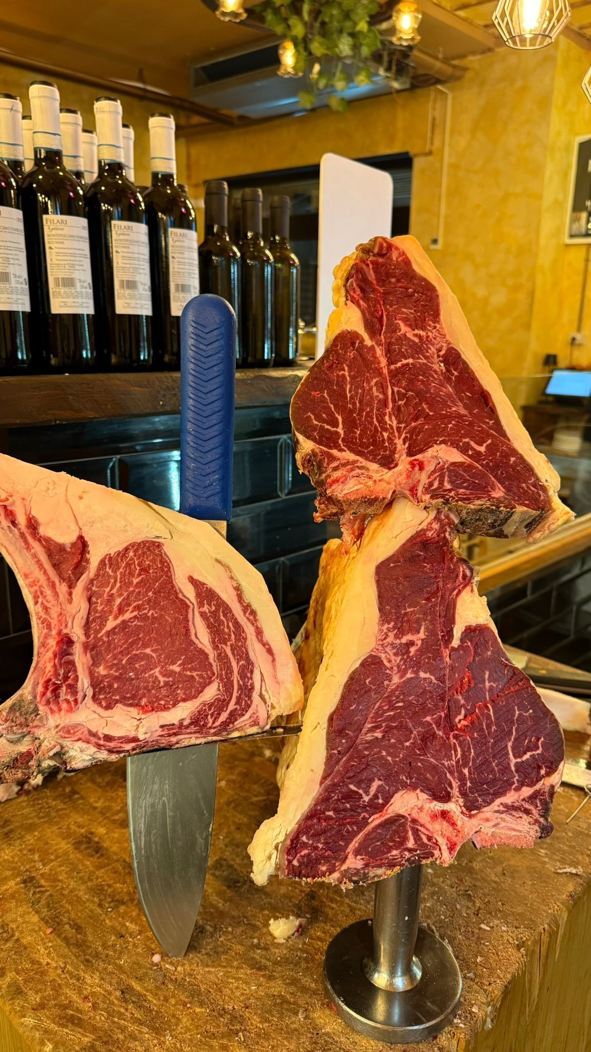 Bistecche crude, tra cui T-bone e ribeye, esposte su un bancone da macelleria, con bottiglie di vino sullo sfondo.