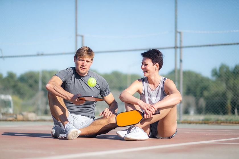 Man and woman sit on a tennis court with pickleball paddles and a ball, smiling.