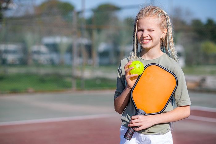 Girl smiles, holding a yellow pickleball and orange paddle on a pickleball court.
