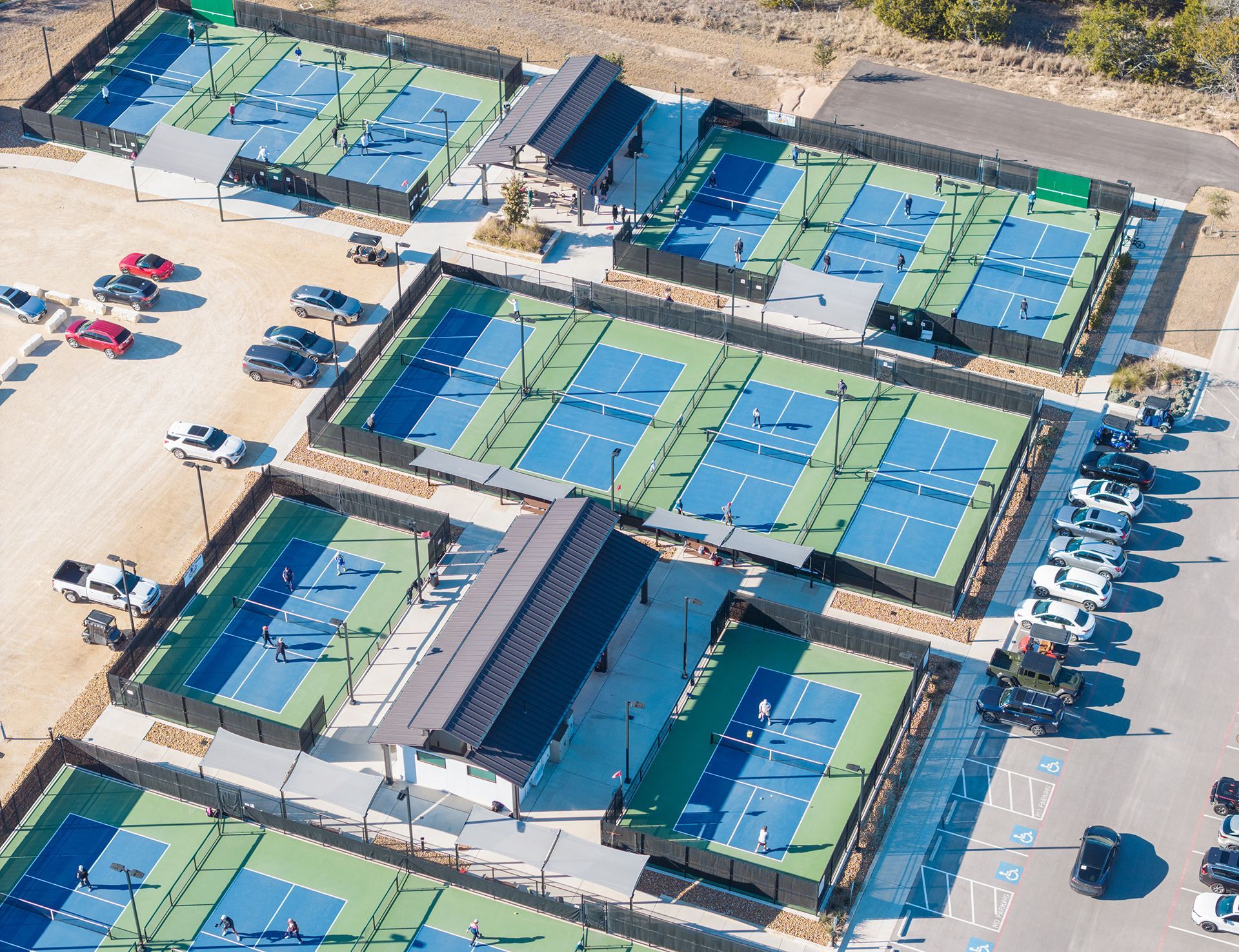 Aerial view of several pickleball courts, blue and green surfaces, with surrounding fencing and parked cars.