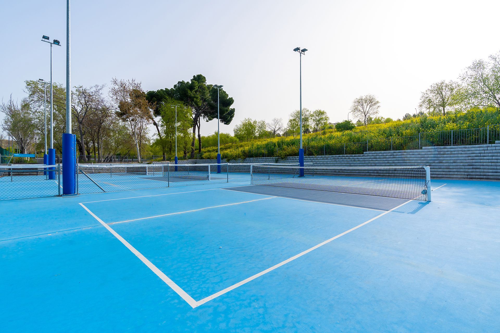 Blue outdoor volleyball court, net, poles, and white lines. Green trees and seating in the background.