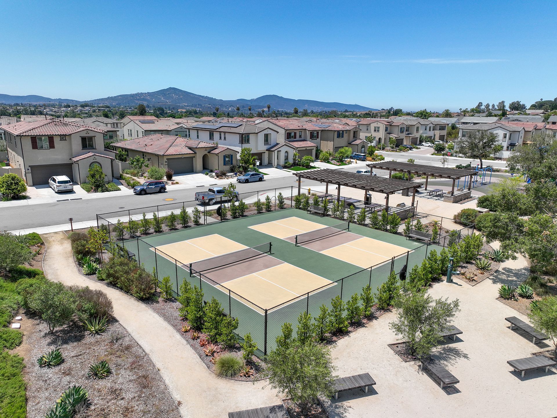 Aerial view: Four pickleball courts surrounded by landscaping, benches, and suburban homes under a blue sky.