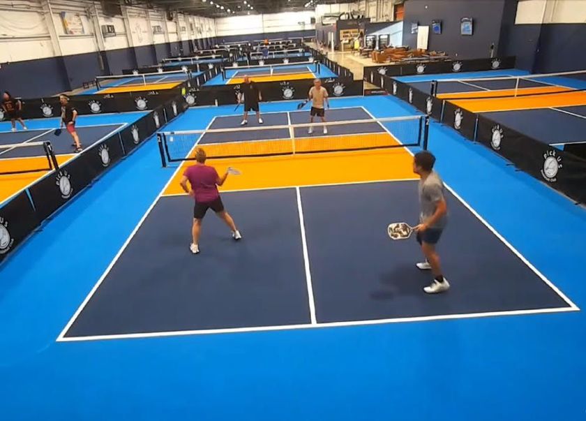 Man and woman sit on a tennis court with pickleball paddles and a ball, smiling.