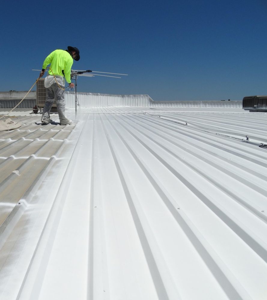 Worker in protective gear spraying white coating on a metal roof under a blue sky.