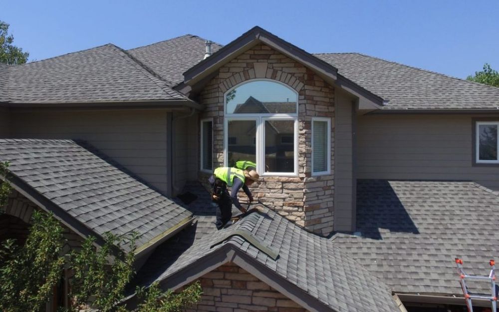 Two workers in safety vests on a rooftop, near a window, with shingles and stone siding.