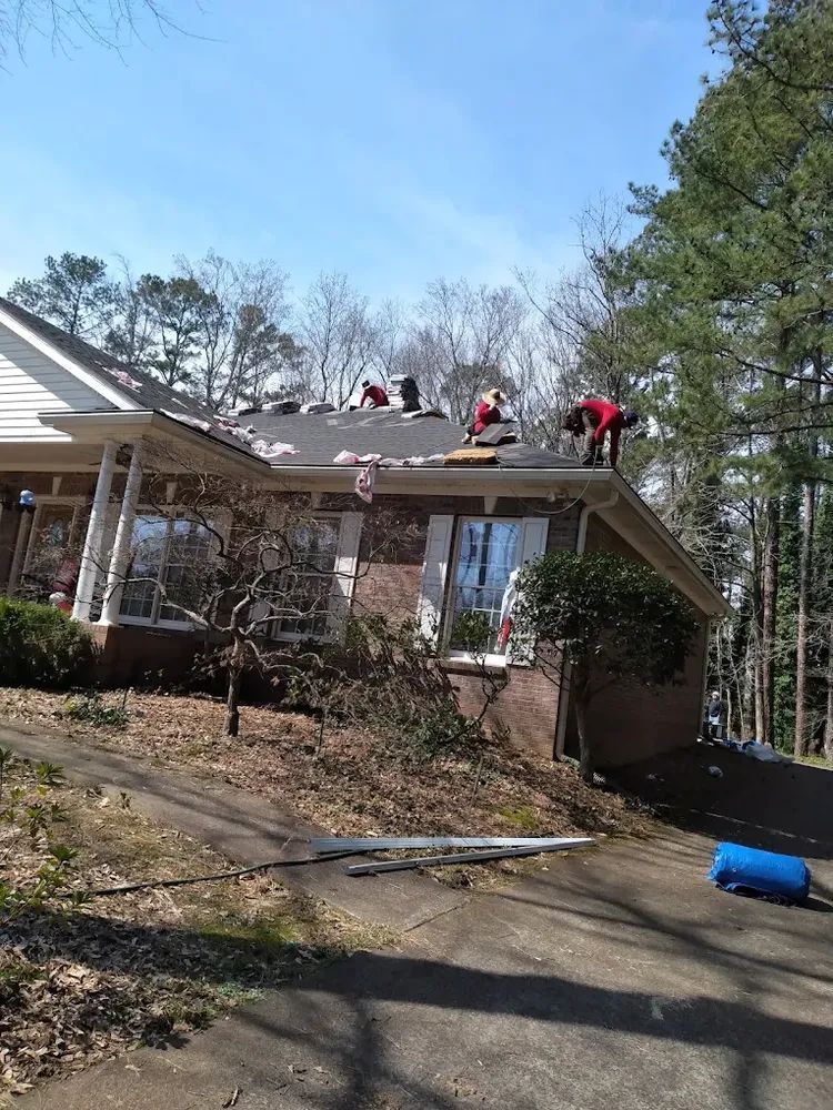 Roofers in red shirts work on a residential roof under a blue sky, trees in background.