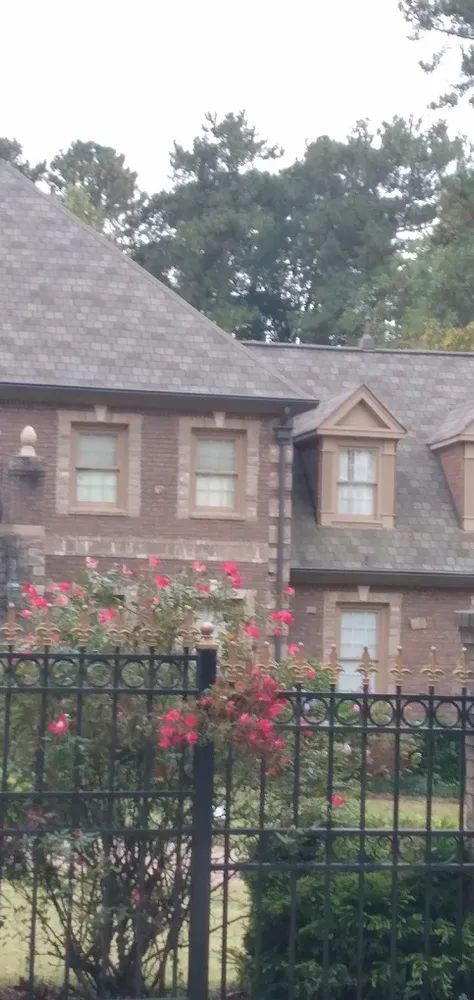 Brick house with multiple windows and a gray roof, behind a black wrought-iron fence.