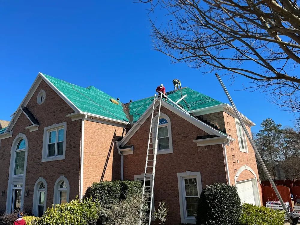 Roofers on a brick house with green shingles, working under a clear blue sky, using ladders.