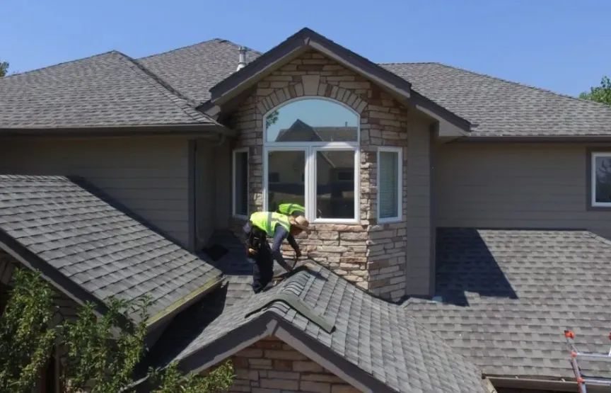 Roofer in safety gear works on the gray shingled roof of a house with stone accents on a sunny day.