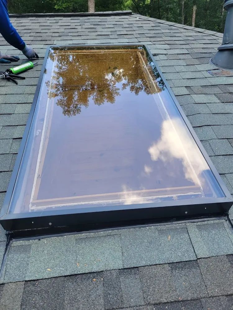 Skylight on a shingled roof, person sealing edges. Trees and sky reflected in the glass.