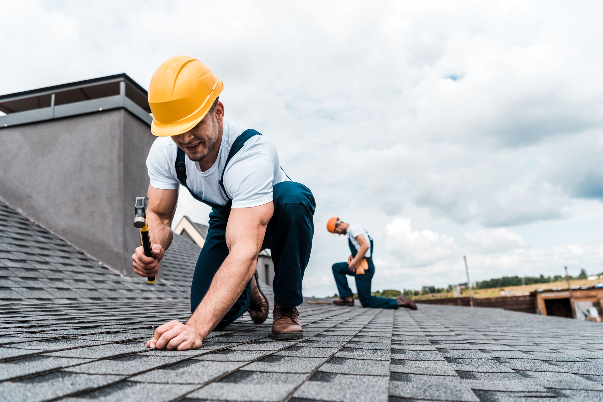 Two roofers in safety gear install shingles on a rooftop under a cloudy sky.