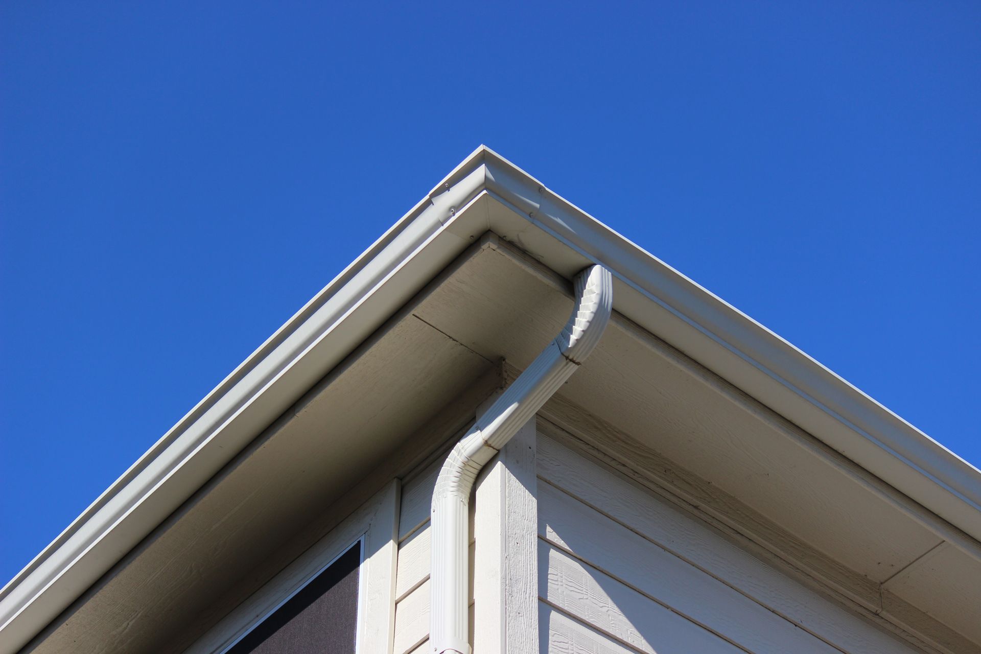 Beige house corner with gutter and downspout against blue sky.