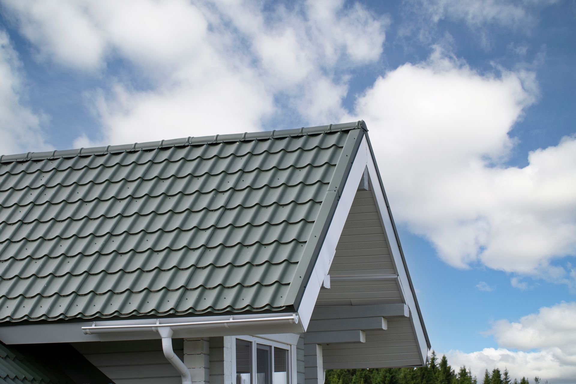 Green tiled roof of a house with white trim against a cloudy blue sky.