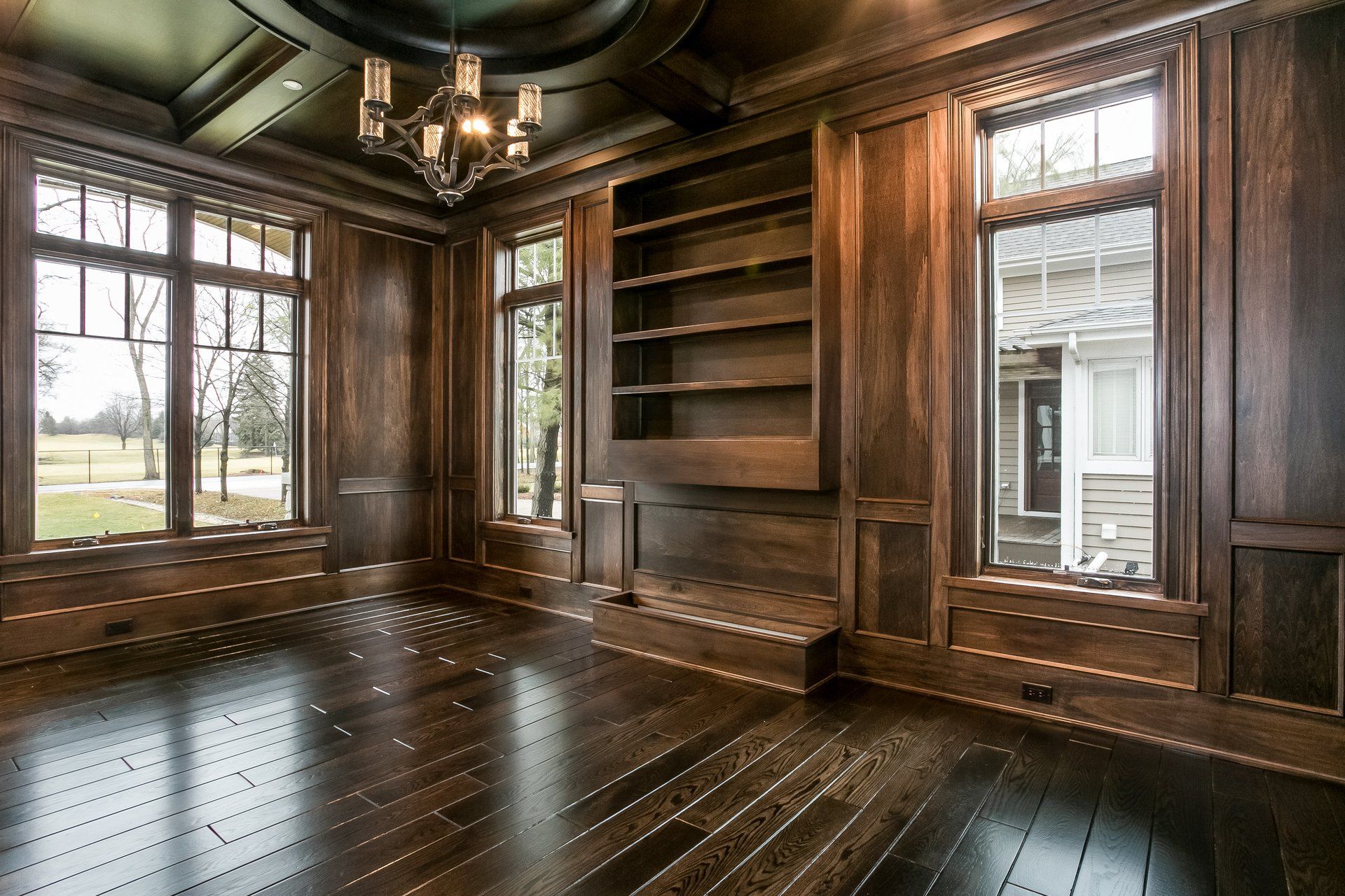 An empty room with wood paneling , hardwood floors , windows and a chandelier.