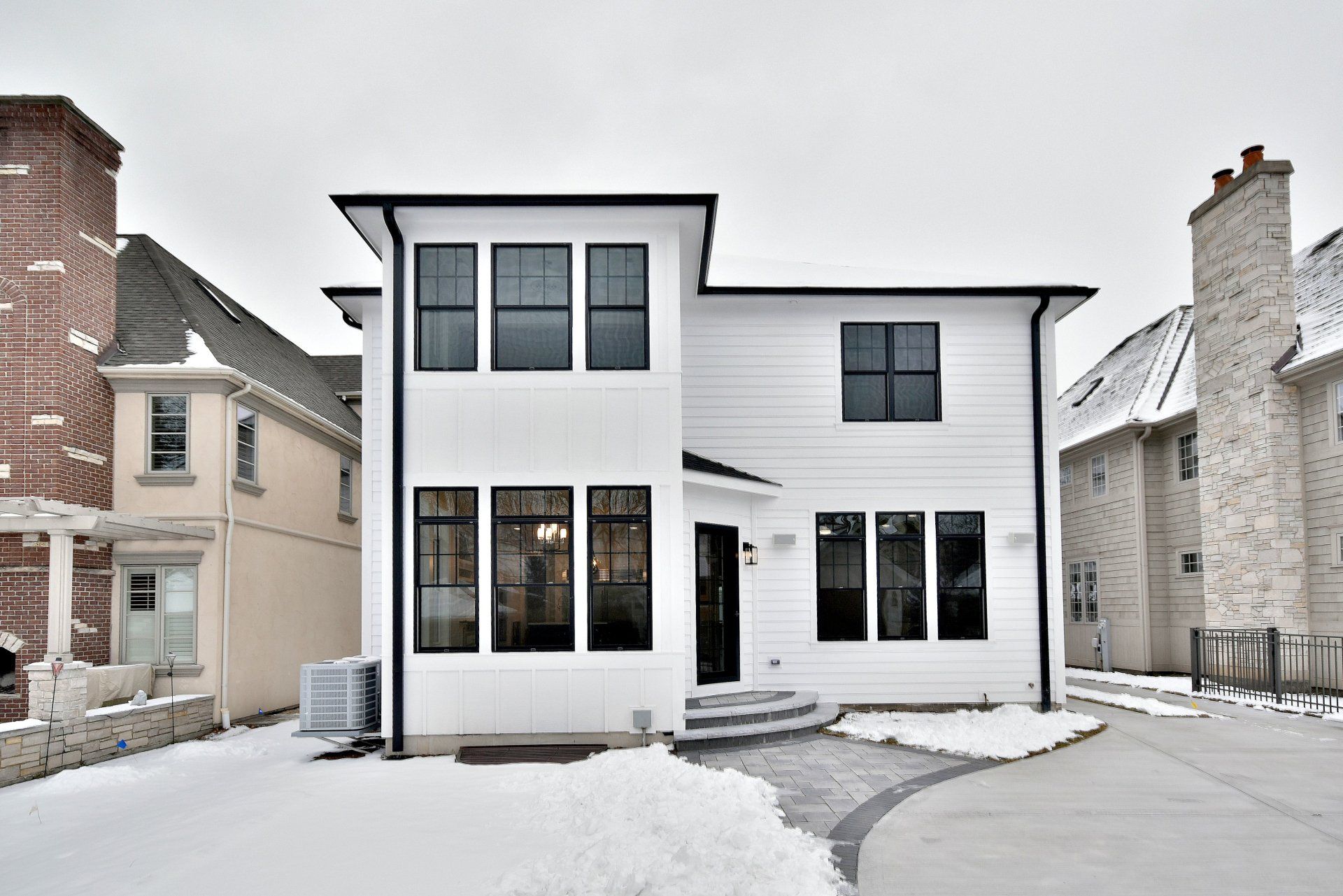 A white house with black windows is surrounded by snow and brick houses.