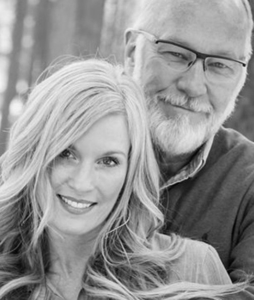 Smiling couple embracing outdoors: blonde woman, older man with glasses.