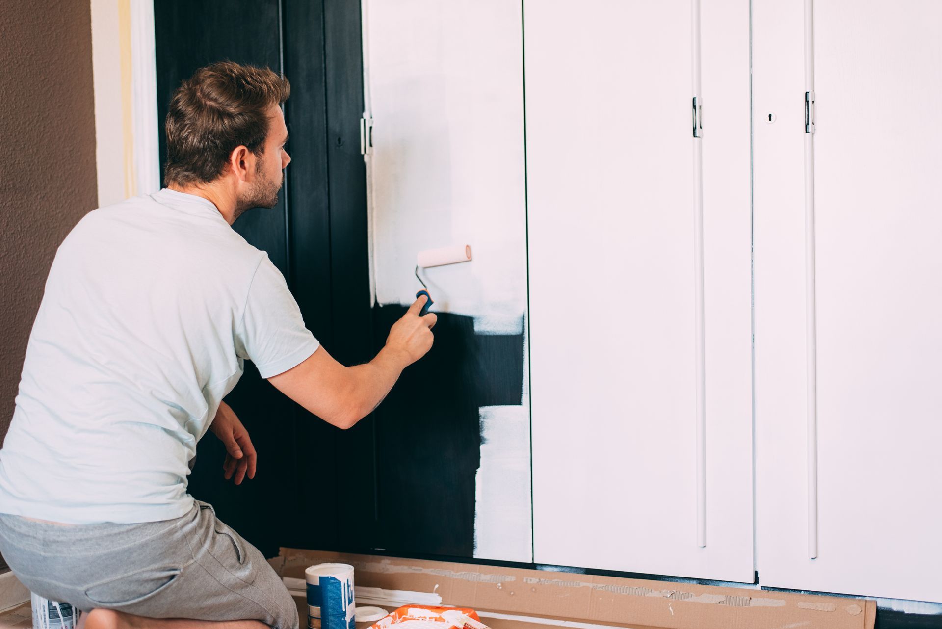 Man paints a black wall with white paint, using a roller; kneeling in front of white closet doors.
