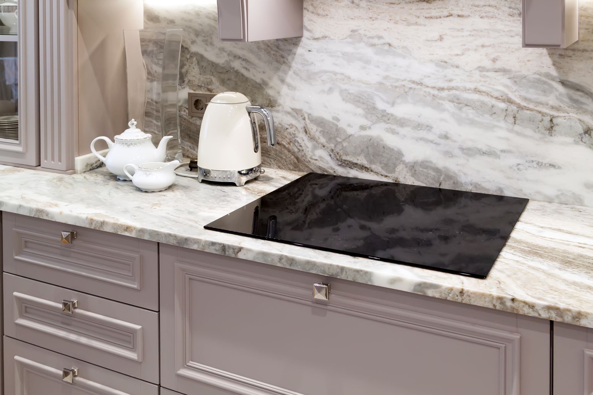 Kitchen countertop with a teapot, kettle, and cooktop. Gray and white marble, gray cabinets.