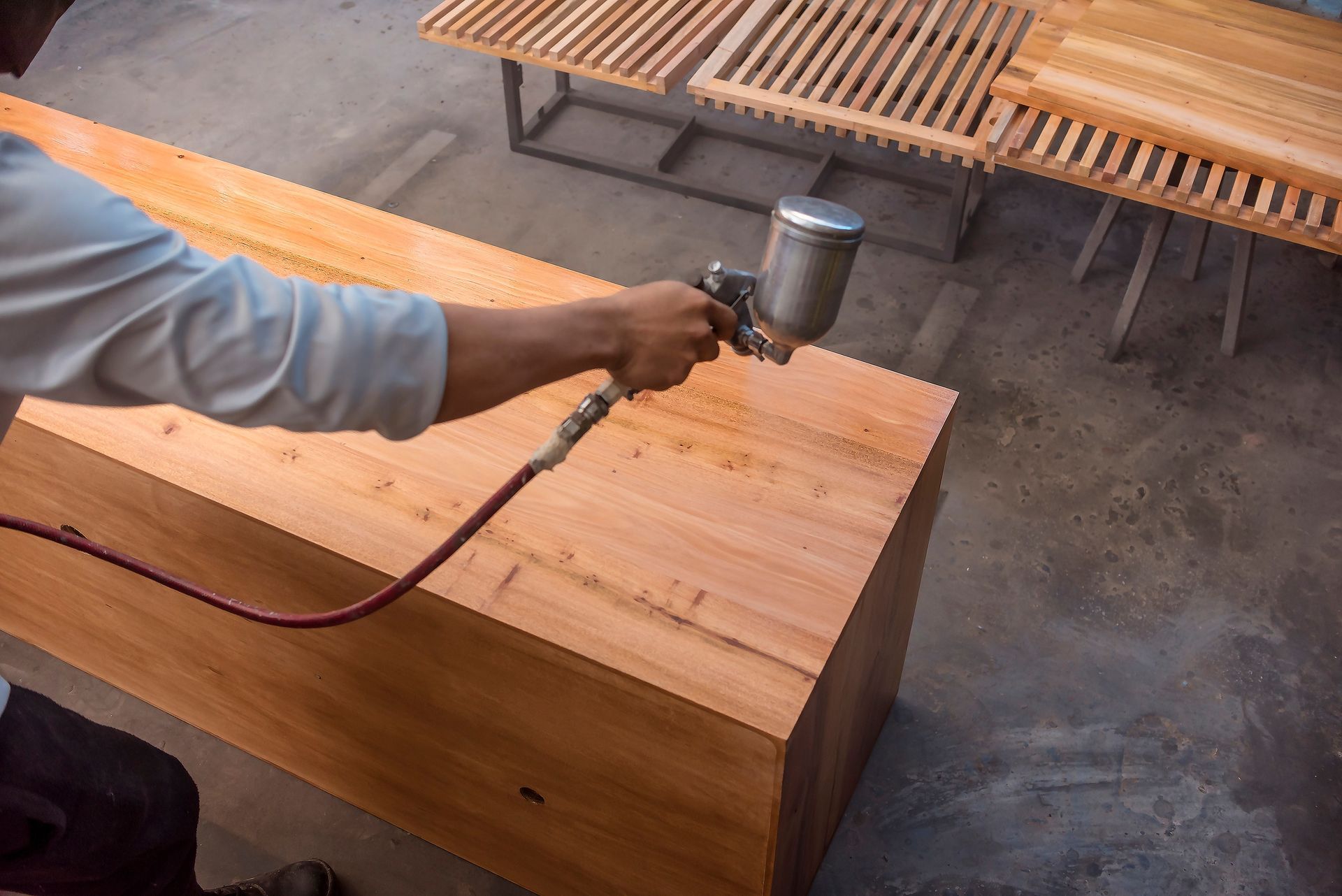 A person uses a spray gun to apply a clear finish to a wooden surface in a workshop.